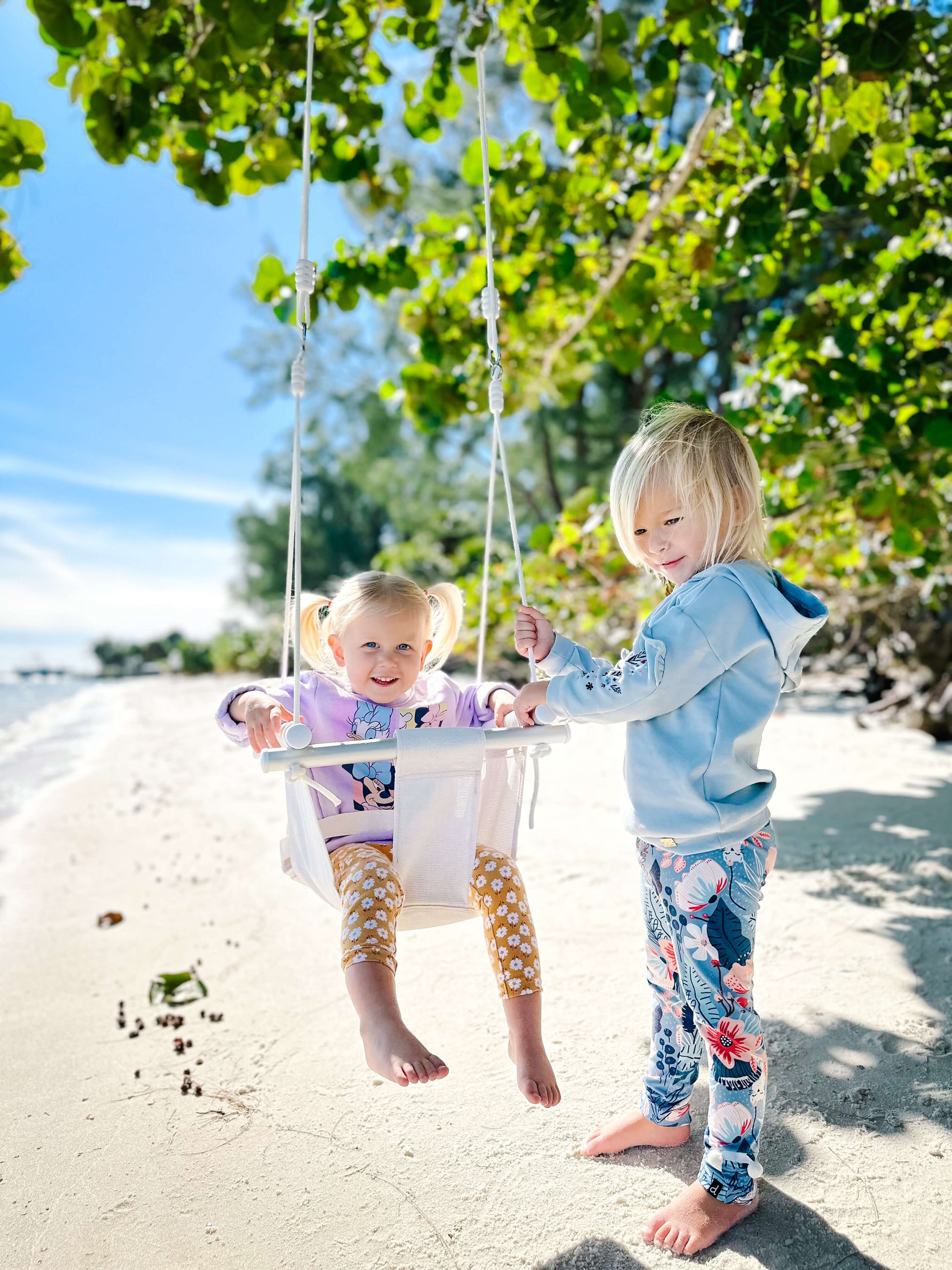 Two children on a beach, one on a Salty Nest Outdoor swing held by the other, with trees and sand in the background.