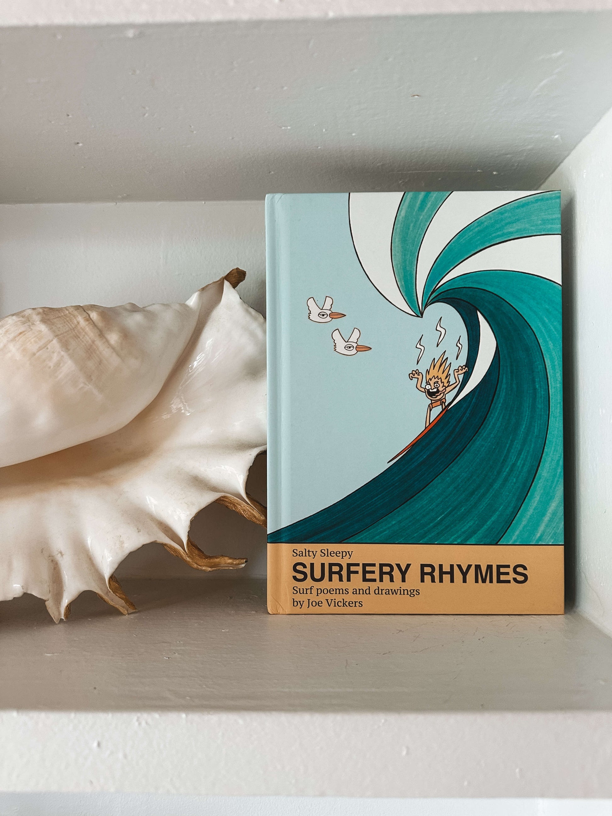 Book titled 'Surfery Rhymes' with a wave design on a shelf next to a shell