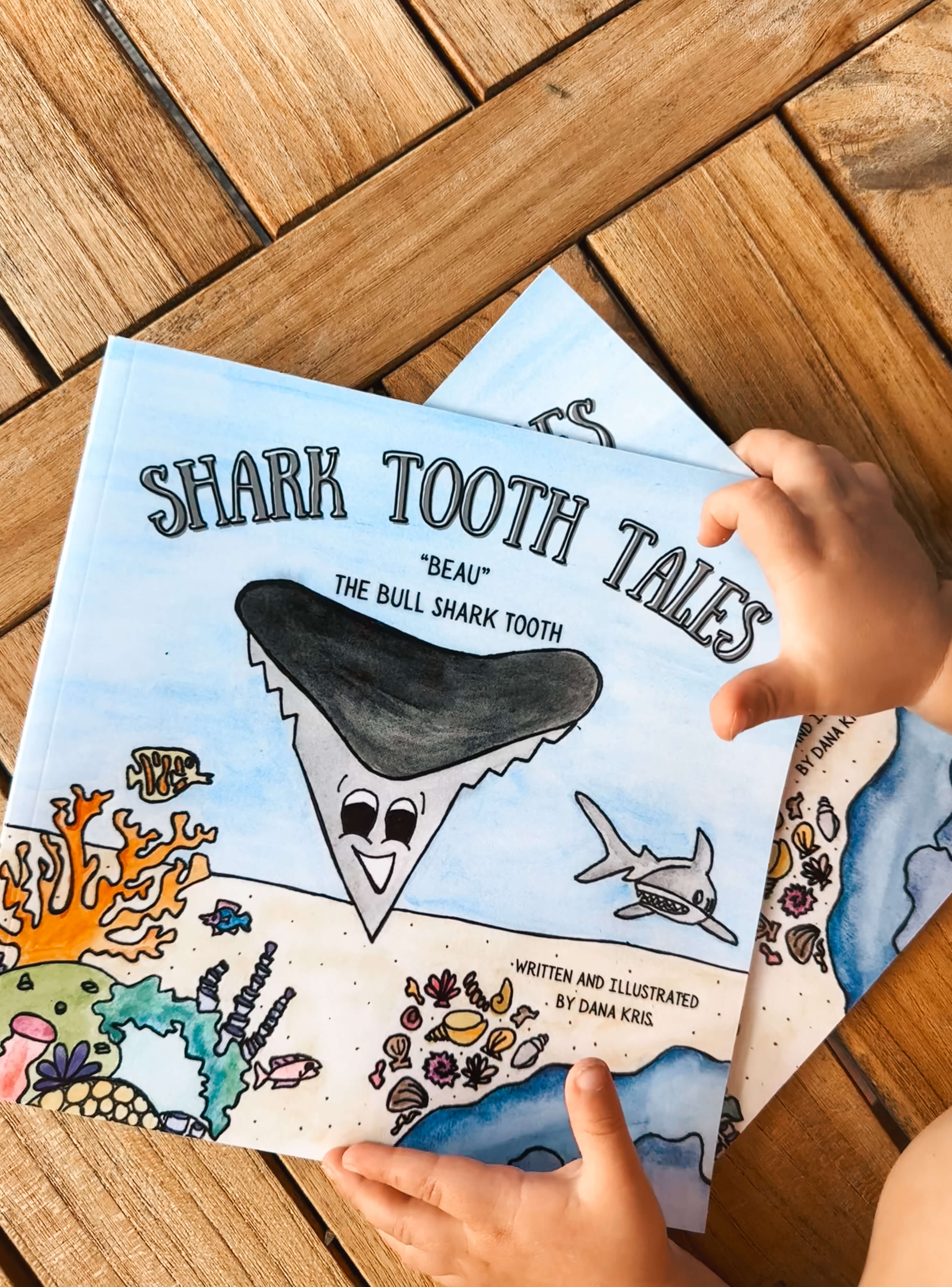 A child is holding up a book titled 'Shark Tooth Tales: The Ball Shark Tooth' by Dana Kris. The cover features colorful illustrations of sea life, including a whale, a crab, and a shark.