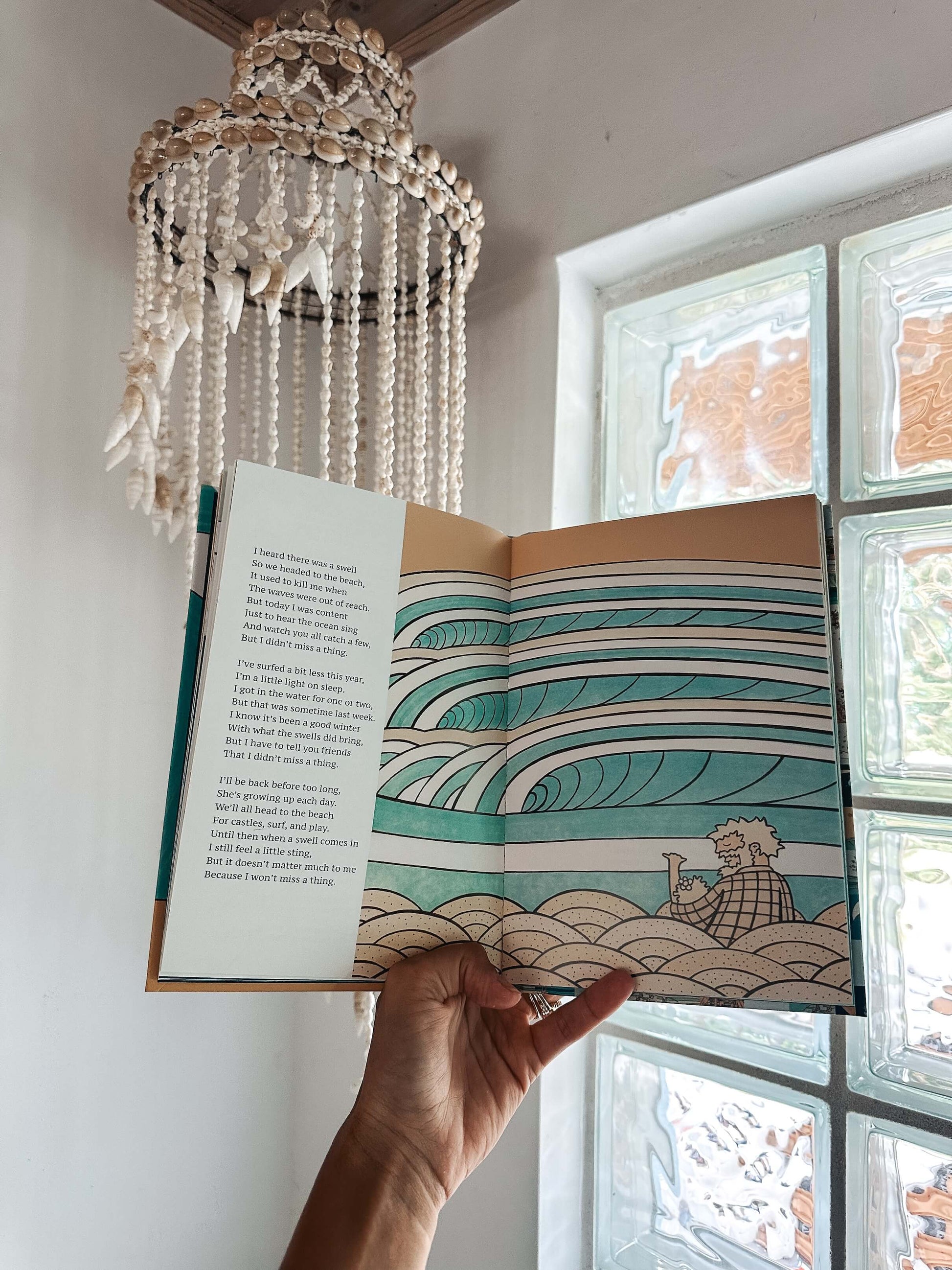 Open book with ocean-themed design held by a hand, with a decorative chandelier and window in the background.