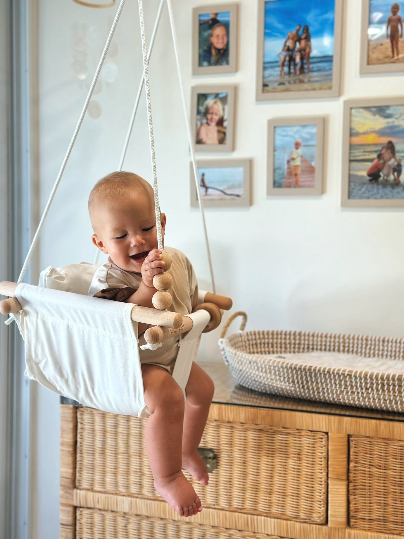 Baby sitting in a white baby Swing surrounded by framed family photos on the wall.